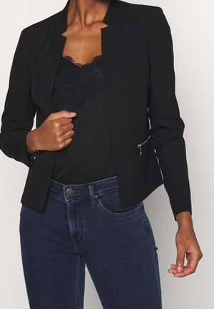 Femme portant un blazer noir par-dessus un haut noir bordé de dentelle, assorti à un jean en denim bleu foncé, tenant le blazer d'une main.
