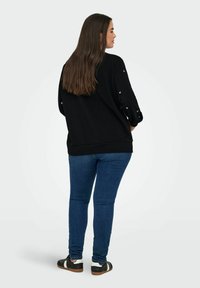 Schwarzer, übergroßer Sweatshirt mit pinken Herzdetails an den Ärmeln, kombiniert mit engen blauen Jeans und schwarzen Sneakers mit weißen Akzenten.