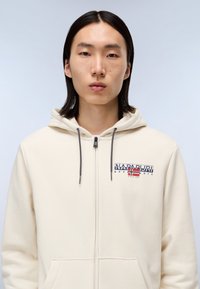 Felpa con zip crema, con lacci grigi, caratterizzata da un patch logo blu e rosso. Tessuto morbido, tasca a canguro e polsini a coste.