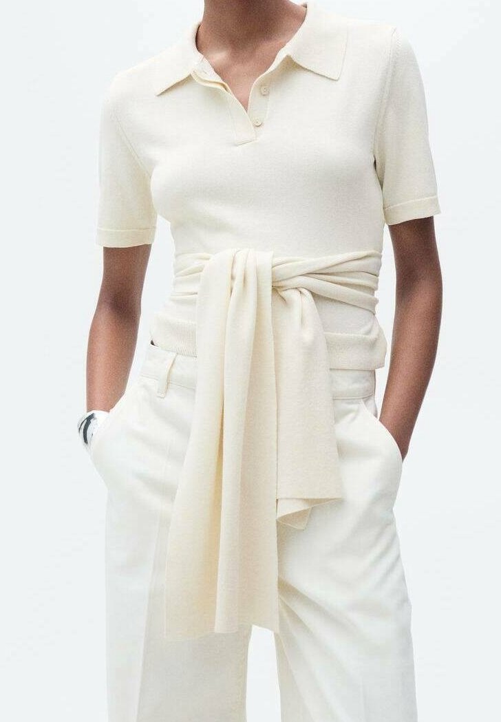 Personne portant un polo crème à manches courtes avec un pull assorti noué autour de la taille et un pantalon blanc, les mains dans les poches.