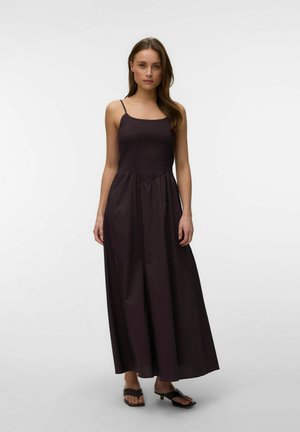 Kvinde iført en sort maxikjole uden ærmer med tynde stropper og sorte kitten heel-sandaler, stående mod en ensfarvet hvid baggrund.