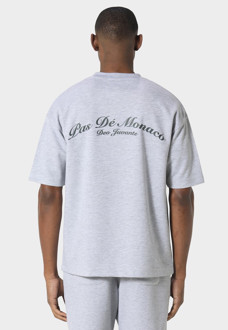 Man draagt lichtgrijs kortärmelig shirt en broek, staat met zijn rug naar de camera, het shirt heeft de tekst "Pas Dé Monaco Deo Juvante."