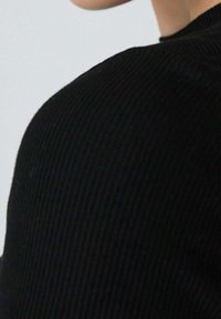 Černý žebrovaný svetr zaměřený na oblast ramena, ukazující strukturovanou tkaninu a bezšvový design. Jemný zakřivený výstřih.