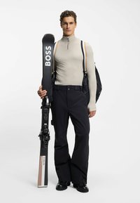 Sprzęt narciarski składający się z dopasowanej beżowej bluzki z długim rękawem, czarnych spodni i czarnych nart z logo "BOSS". Trzyma narty i nosi czarny plecak.