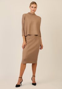 Hellbraunes Strick-Set, bestehend aus einem kurzen, weiten Pullover mit gerippten Ärmeln und einem figurbetonten Midirock. Kombiniert mit schwarzen offenen Zehen-Pumps.