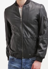 Giubbotto bomber in pelle nera con finitura testurizzata, colletto e polsini a coste, zip frontale e tasche laterali, indossato sopra una camicia chiara.