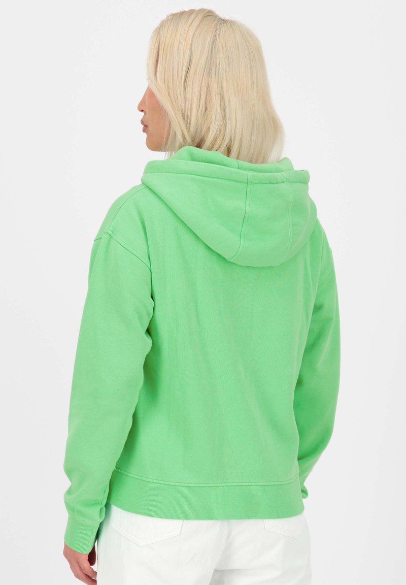 Un sweat à capuche vert en tissu doux, avec une capuche à cordon, des poignets et un ourlet côtelés, et une coupe décontractée. Des pantalons blancs sont partiellement visibles.