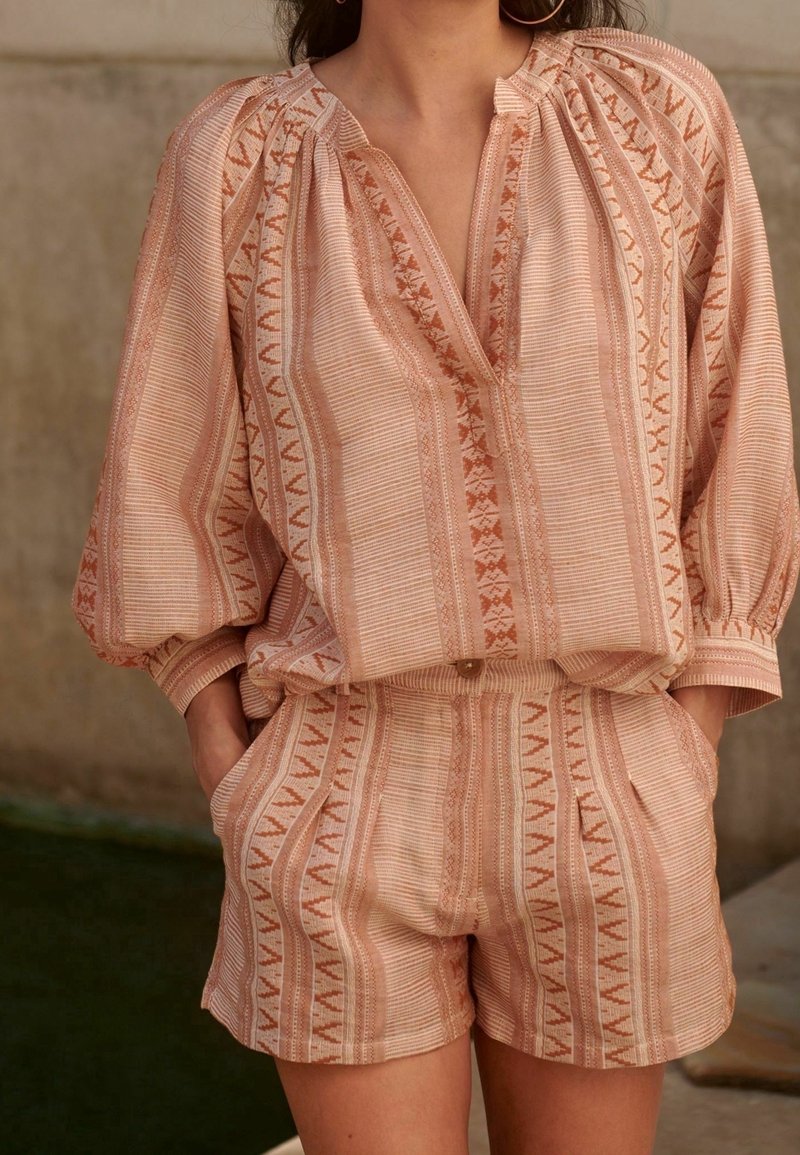 Femme portant un chemisier à manches longues à motifs rose clair et beige avec un short taille haute assorti, mains dans les poches, sur fond neutre.