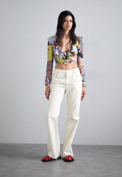 Värikäs leikattu pusero rypytetyn kaula-aukon ja kuviollisten hihojen kanssa; yhdistettynä vaalean beigeihin leveälahkeisiin housuihin ja punaisiin sandaaleihin, joissa on musta pohja.