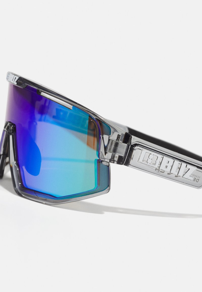 Sonnenbrille mit klarem Rahmen, großen Blaufarben und lila Farbverlauf-Gläsern, kantigem Design und auffälligem Branding an den Bügeln.