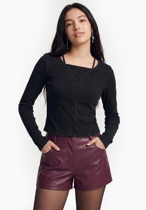 Nuori nainen, jolla on pitkät tummat hiukset, pukeutuneena mustaan napitettuun yläosaan ja burgundin punaisiin nahkashortseihin, seisoo käsivarret taskuissa valkoista taustaa vasten.