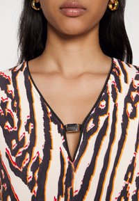 Femme portant un haut à motifs abstraits multicolores avec un col en V et un détail de fermoir métallique, ainsi que des boucles d’oreilles créoles dorées.