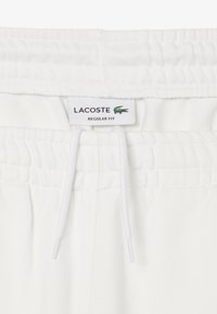 Pantalones de chándal blancos de corte regular con cinturilla elástica, cordón y etiqueta de Lacoste. Textura suave con sutiles detalles de costura.