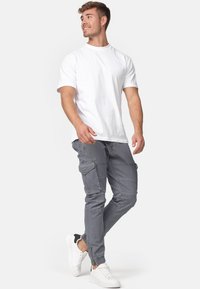 Fiatal férfi sétál, sima fehér pólót, szürke cargo nadrágot és fekete részletekkel ellátott fehér sportcipőt visel, oldalra néz és mosolyog.