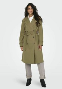 Olivgrön trench coat med dubbelknäppt design, bälte i midjan, stora kragar och sidofickor, parat med ljusgrå uppvikta byxor och svarta stövlar.