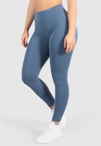 Leggings a vita alta e aderenti in un morbido tessuto blu, caratterizzati da una superficie liscia e cuciture minime, abbinati a scarpe da ginnastica bianche.