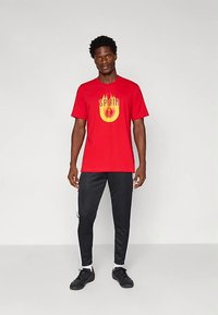 Punainen T-paita, jossa on keltainen graafinen kuva ja sana "SPAIN" sekä liekkikoristeita. Yhdistetty mustiin housuihin, jotka ovat urheilullisen malliset ja istuvat.