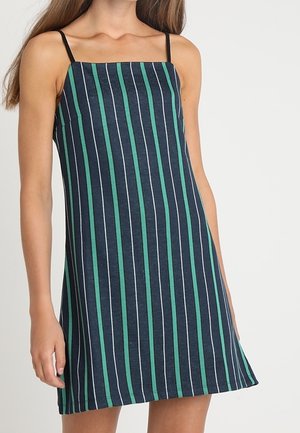 Hihaton laivastonsininen mekko, jossa ohuet mustat olkaimet ja pystysuuntaiset vihreät ja valkoiset raidat, päällä seisovalla henkilöllä yksivärisen taustan edessä.