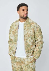 Homme avec des cheveux bouclés courts portant une veste à motif floral et un pantalon assorti, les mains dans les poches, regardant sur le côté.