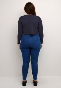 Kurz geschnittenes navyblaues Langarmoberteil, kombiniert mit blauen hochgelegten Skinny-Jeans. Die Jeans verfügen über Gesäßtaschen und haben einen figurschmeichelnden Schnitt. Schwarze Schuhe runden den Look ab.