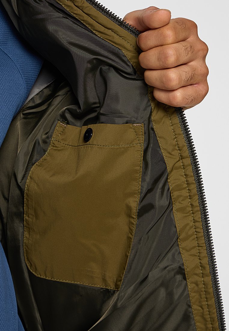 Olivgrünes Innenfach mit Knopfverschluss, umgeben von glattem, dunkelgrauem Stofffutter innerhalb einer Jacke. Sichtbarer Reißverschlussdetails.