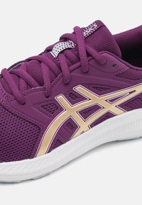 Chaussure de sport violette avec une tige en maille, des accents dorés, des lacets texturés et une semelle en caoutchouc blanc avec des détails en motif.