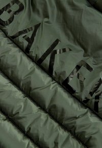 Giacca a vento verde oliva con grandi lettere nere che scrivono "BAKER" sulla parte frontale, caratterizzata da una texture liscia e lucida e un design trapuntato.