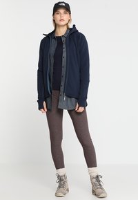 Sininen vetoketjullinen fleece-takki tumman paidan, harmaan nappipaidan, ruskeiden leggingsien ja vaaleanharmaiden neulekoristeilla varustettujen saappaiden päällä. Musta lippalakki.