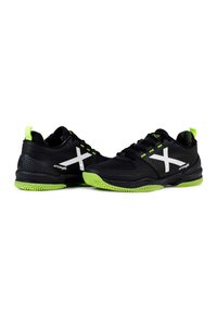 Zapatillas deportivas negras con un logo blanco en forma de "X", detalles verdes en el talón y la suela, con un upper texturizado y diseño transpirable.