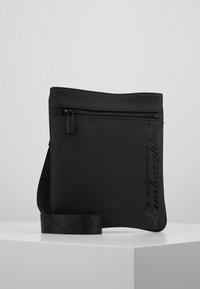 Černá ramenní taška vyrobená z texturovaného materiálu. Má horní zip a vzorovaný popruh pro snadné nošení. Ozdobena raženým textem.