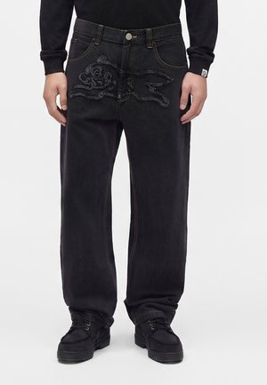 Jean noir ample avec un motif texturé d’un lapin courant sur le devant, porté avec des chaussures noires et un haut noir à manches longues.