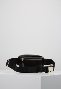 Černá kožená pásková taška s přední zipovou kapsou a nastavitelným popruhem se zlatou kovovou přezkou na bílé ploše.