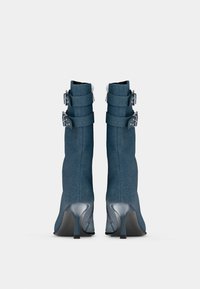 Denim gležnjarji s tankim stiletto peto, ki vključujejo tri dekorativne kovinske zaponke in teksturirano tkanino v temno modri barvi.