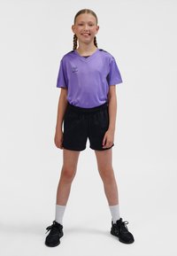 Maglietta sportiva viola con dettagli neri, abbinata a pantaloncini neri. L'outfit presenta una vestibilità rilassata ed è completato da calzini bianchi a costine e scarpe da ginnastica nere.