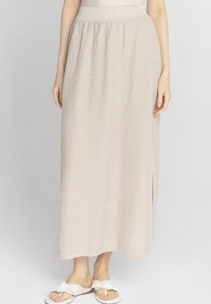 Kobieta ubrana w beżową, teksturowaną spódnicę midi z wysokim stanem i bocznym rozcięciem, połączoną z białymi sandałami typu thong z węzłami.