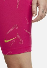 Rožinės sportinės šortai su pasikartojančiu šviesios aukso spalvos Nike logotipo raštu. Audinys atrodo lygus, dizainas prigludęs.