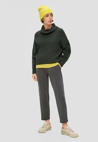 Grüner Strickpullover mit einem Schalkragen, über einem gelben Oberteil getragen, kombiniert mit grauen Hosen und beigen Sneakers. Gelbe Strickmütze als Accessoire.