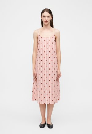 Giovane donna in piedi che indossa un vestito midi senza maniche a righe rosa e bianche con piccoli motivi floreali rossi e scarpe nere basse.