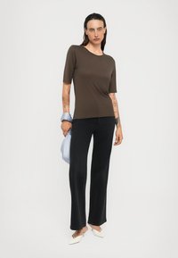Donna con top a maniche corte color marrone scuro, jeans neri a gamba larga, tacchi bianchi a punta, che tiene una borsa azzurro chiaro su uno sfondo bianco.