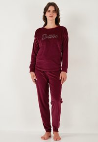 Burgundski velur pajama komplet z dolgimi rokavi in ujemajočimi se hlačami. Vsebuje besedilo "sledi svojim sanjam" in mehko, teksturirano tkanino.