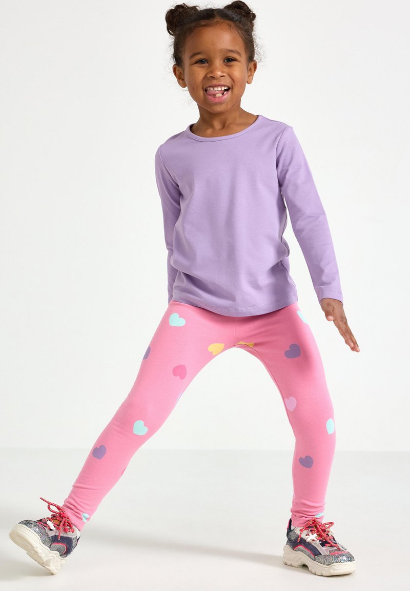 Długi rękaw w kolorze bzu połączony z różowymi legginsami z wielokolorowymi wzorami serc. Do tego wielokolorowe sneakersy, na jednolitym tle.