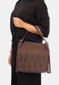 Bolso de ante marrón con detalles de flecos, una superficie texturizada y una única correa para el hombro. El bolso tiene un diseño estructurado con forma redondeada.