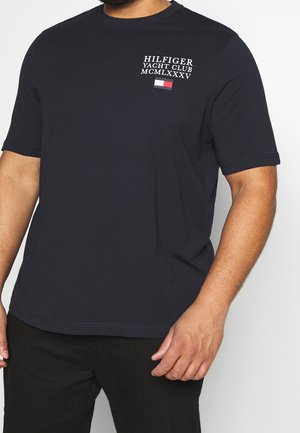 Mand iført en navyblå T-shirt med korte ærmer med teksten "Hilfiger Yacht Club MCMLXXXV" i hvid samt et rødt, hvidt og blåt logo på venstre bryst.