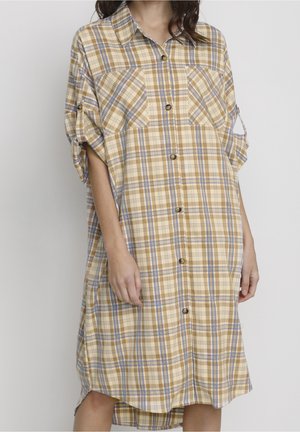 Kobieta ubrana w beżową, niebieską i białą sukienkę w kratę sięgającą do kolan, z podwiniętymi rękawami i dwoma kieszeniami na piersiach.
