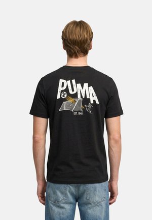 Uomo che indossa una maglietta nera PUMA con grafica di una porta da calcio e palla che entra in rete, in piedi davanti a uno sfondo semplice.