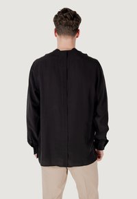 Camicetta nera a maniche lunghe con taglio dritto, cucitura posteriore sottile e polsini con bottoni, abbinata a pantaloni beige chiari. Tessuto dalla texture liscia.