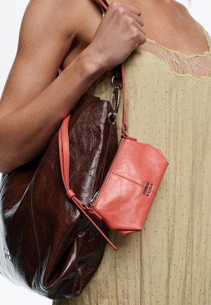 Ręka trzymająca dwie skórzane torebki, jedną dużą w ciemnym brązie i jedną małą w koralowym różu, przewieszone przez ramię, ubraną w jasnżółtą sukienkę z koronkowym wykończeniem.