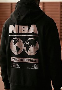 Personne portant un sweat à capuche noir avec un grand texte "NBA", deux illustrations de globe et le slogan "Portail vers le Rêve" au dos.