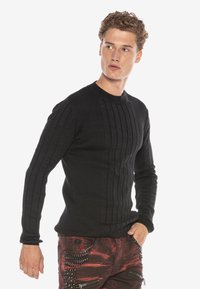 Schwarzer, langärmliger Ripp-Pullover mit senkrechten Mustern, Rundhalsausschnitt und figurnahem Schnitt, getragen mit roten, distressten Hosen mit Reißverschlüssen.