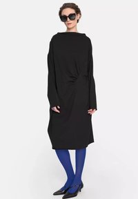 Czarna sukienka z długim rękawem, okrągłym dekoltem i węzłem w talii. Noszona z niebieskimi rajstopami i czarnymi szpilkami.